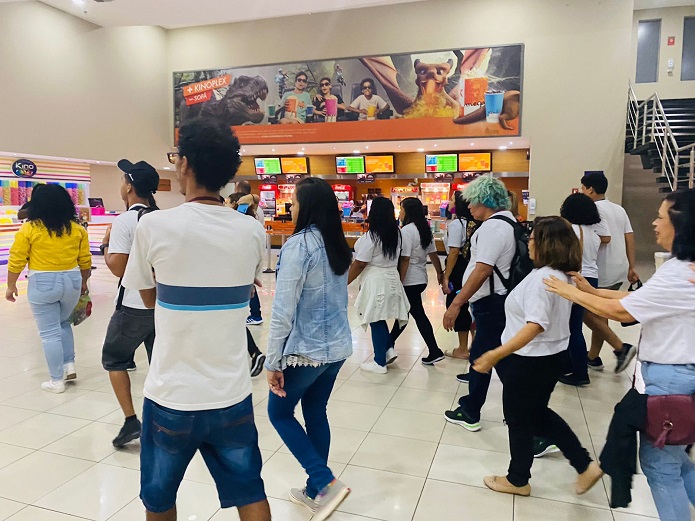 Foto. Grupo de estudantes da Educação de Jovens e Adultos caminha no saguão do cinema. Ao fundo, a lanchonete do cinema, que possui na parede acima um cartaz com imagens de filmes e pessoas com óculos 3D e pacote grande de pipoca nas mãos.