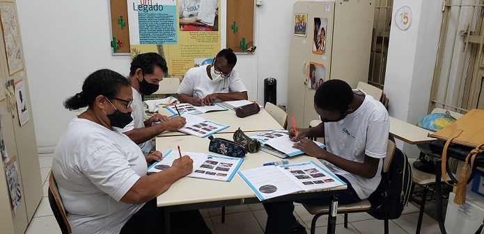 Foto. Quatro alunos escrevem em apostilas, sentados ao redor de uma mesa. Há também sobre a mesa, estojos escolares. Os quatro são negros. Da esquerda para a direita, há uma mulher de meia idade acima do peso, usando óculos e os cabelos presos. A seguir: um homem jovem, um rapaz usando óculos e outro rapaz. Todos usam blusa branca do uniforme escolar. Atrás deles, há dois armários e um quadro de cortiça com cartazes.