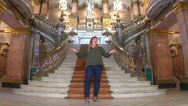 Fotografia colorida que mostra o hall de entrada do Theatro Municipal do Rio, ao centro uma escadaria com corrimão e adornos de época dourados e tapete marrom. No alto, aparece parte dos candelabros. No centro da imagem, professora do Rioeduca na TV em pé com os braços abertos em saudação de boas-vindas.