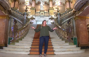 Fotografia colorida que mostra o hall de entrada do Theatro Municipal do Rio, ao centro uma escadaria com corrimão e adornos de época dourados e tapete marrom. No alto, aparece parte dos candelabros. No centro da imagem, professora do Rioeduca na TV em pé com os braços abertos em saudação de boas-vindas.