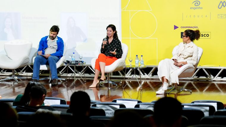 Foto. Três pessoas sentadas em poltronas brancas em cima de um palco.
