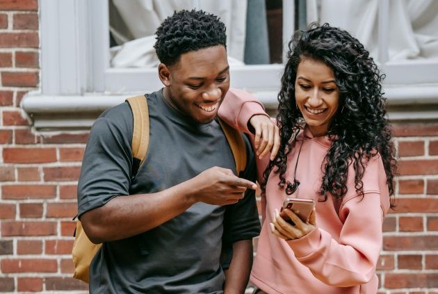 Foto. Uma jovem negra está com um celular em uma mão e a outra apoiada sobre o ombro de um rapaz negro. Ambos sorriem e olham para a tela do celular.egra