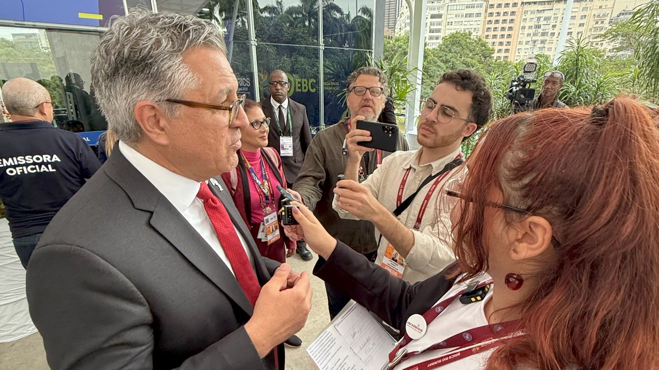 Foto. Homem, de cabelos grisalhos e óculos de grau, vestindo um terno, é entrevistado por um grupo de pessoas, dentre elas uma estudante adolescente.
