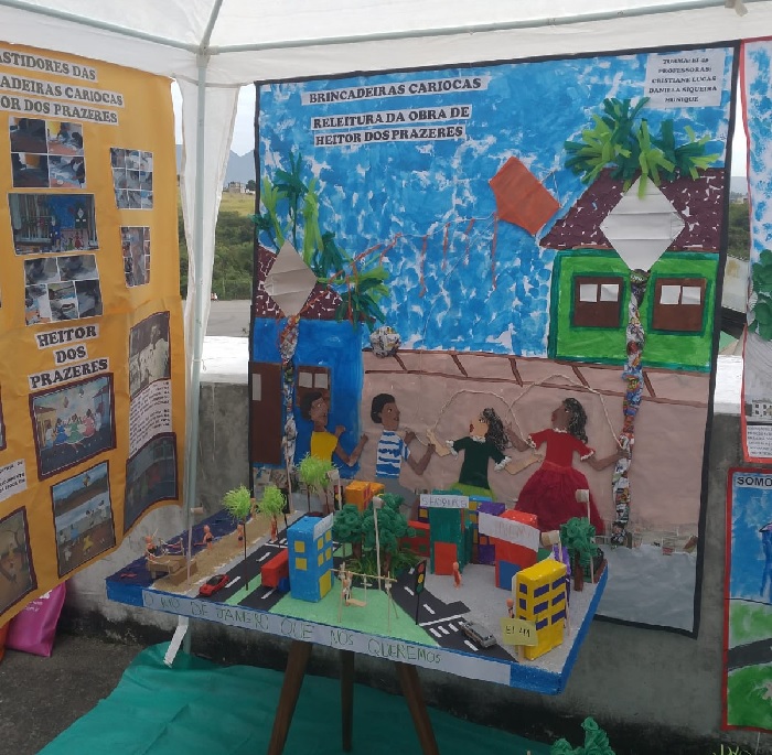 Estande da Creche Luiza Barros na Semana de Educação Infantil com maquete de cidade ao centro e, ao redor, cartazes com fotos, desenhos e colagens