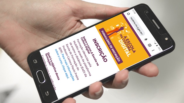 Fotografia colorida que mostra mão segurando um celular em cuja tela aparece a página de inscrição online do curso A Escola na Cultura Digital 