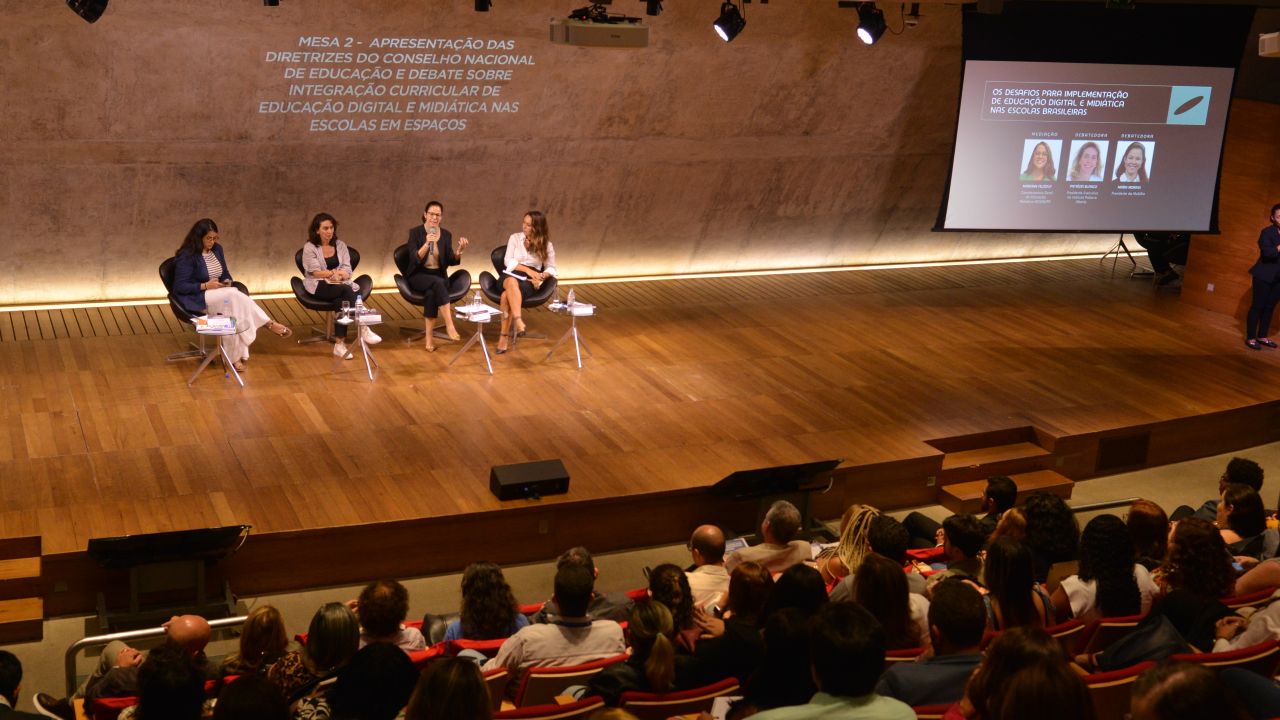 Foto. Quatro mulheres sentadas em cadeiras em um palco. Plateia na parte inferior. A direita, telão com projeção da apresentação.