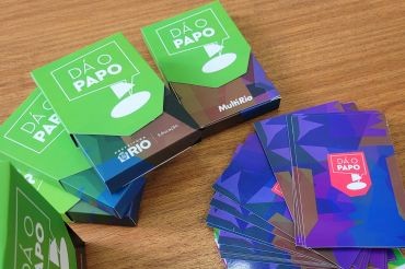 Foto. Caixas e cartas do jogo Dá o Papo estão dispostos sobre uma superfície de madeira. As caixas possuem om logotipo da MultiRio e da Prefeitura do Rio/ Educação.